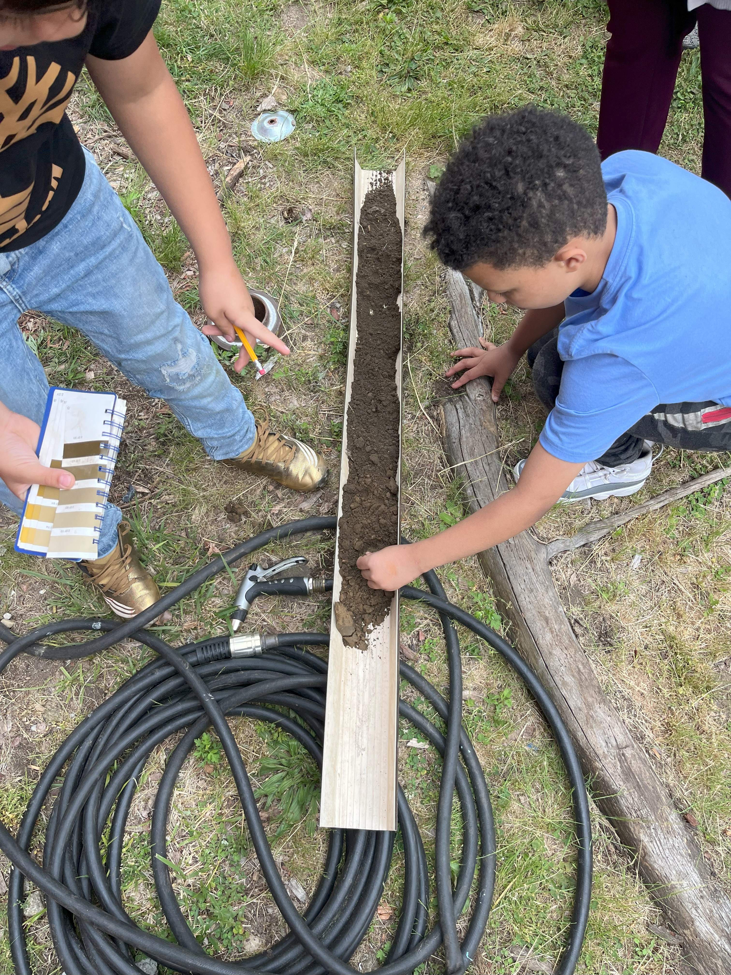 a student crouches on the ground to feel the texture of a soil sample that has been placed in a trough